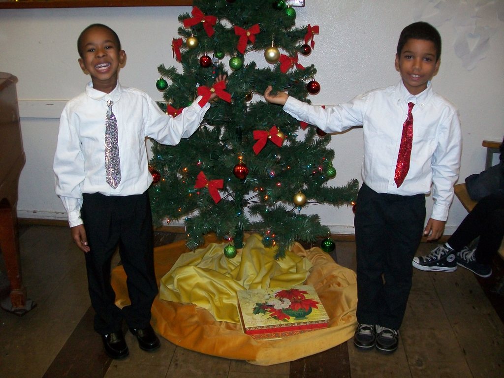 Ein geschmückter Weihnachtsbaum steht in einem Raum mit einem hölzernen Tisch auf der linken Seite, einem Stuhl auf der rechten Seite und zwei Jungen in weißen Hemden, die strahlend den Baum halten, mit verschiedenen Gegenständen und einer Schachtel auf dem Boden und an der Wand im Hintergrund.