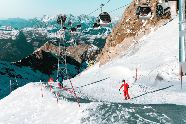 Gruppe von Skifahrern, die eine schneebedeckte Abfahrt hinunterfahren, mit einem Seilbahnwagen im Hintergrund, der mit Gondeln gefüllt ist, vor einer Bergkulisse und einem klaren blauen Himmel; der Skifahrer im Vordergrund trägt eine rote Ausrüstung und hält Ski-Stöcke.
