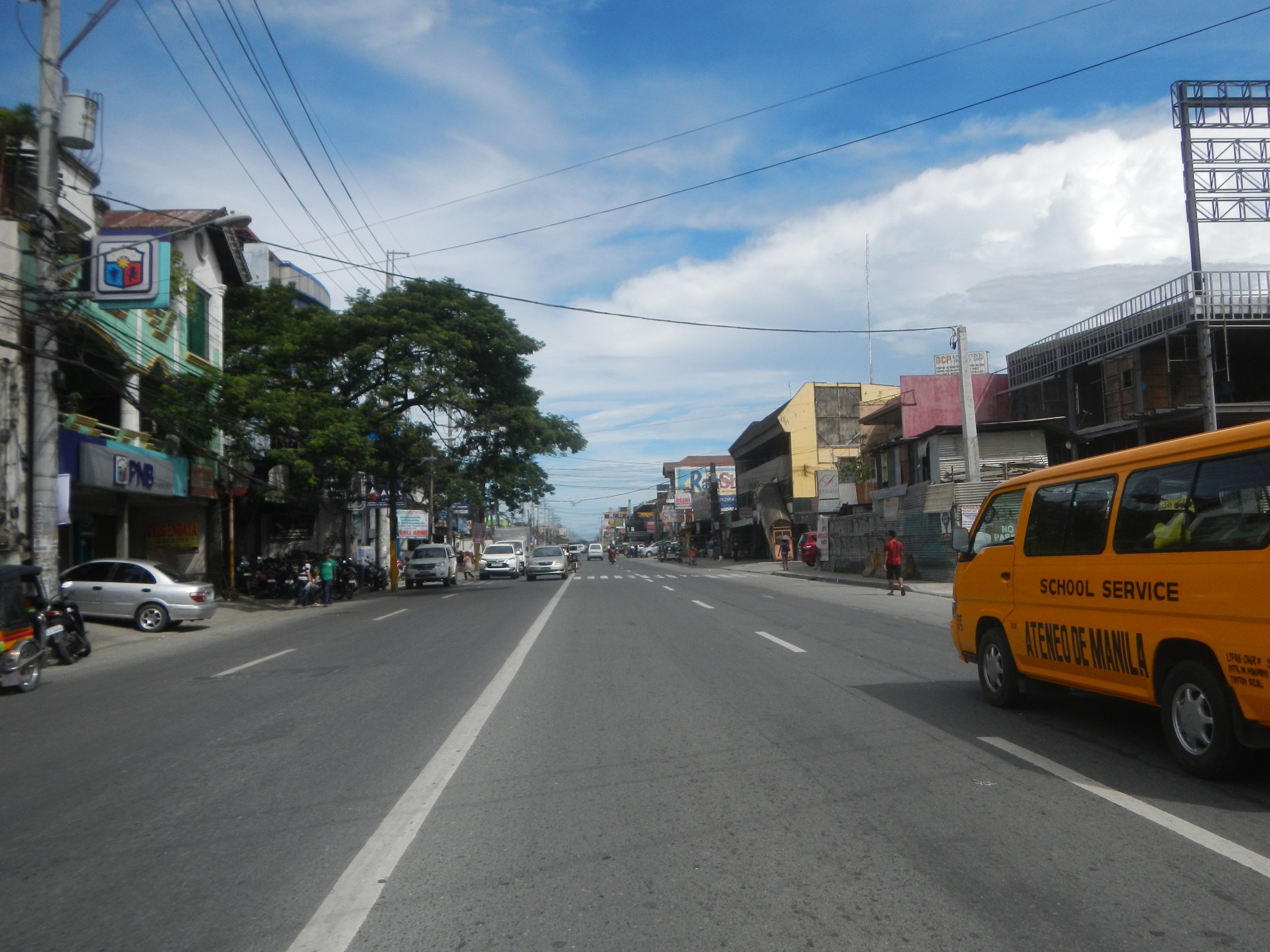 Gelber Schulbus fährt auf einer Straße mit hohen Gebäuden, Passanten, Bäumen, Strommasten und Überlandleitungen.