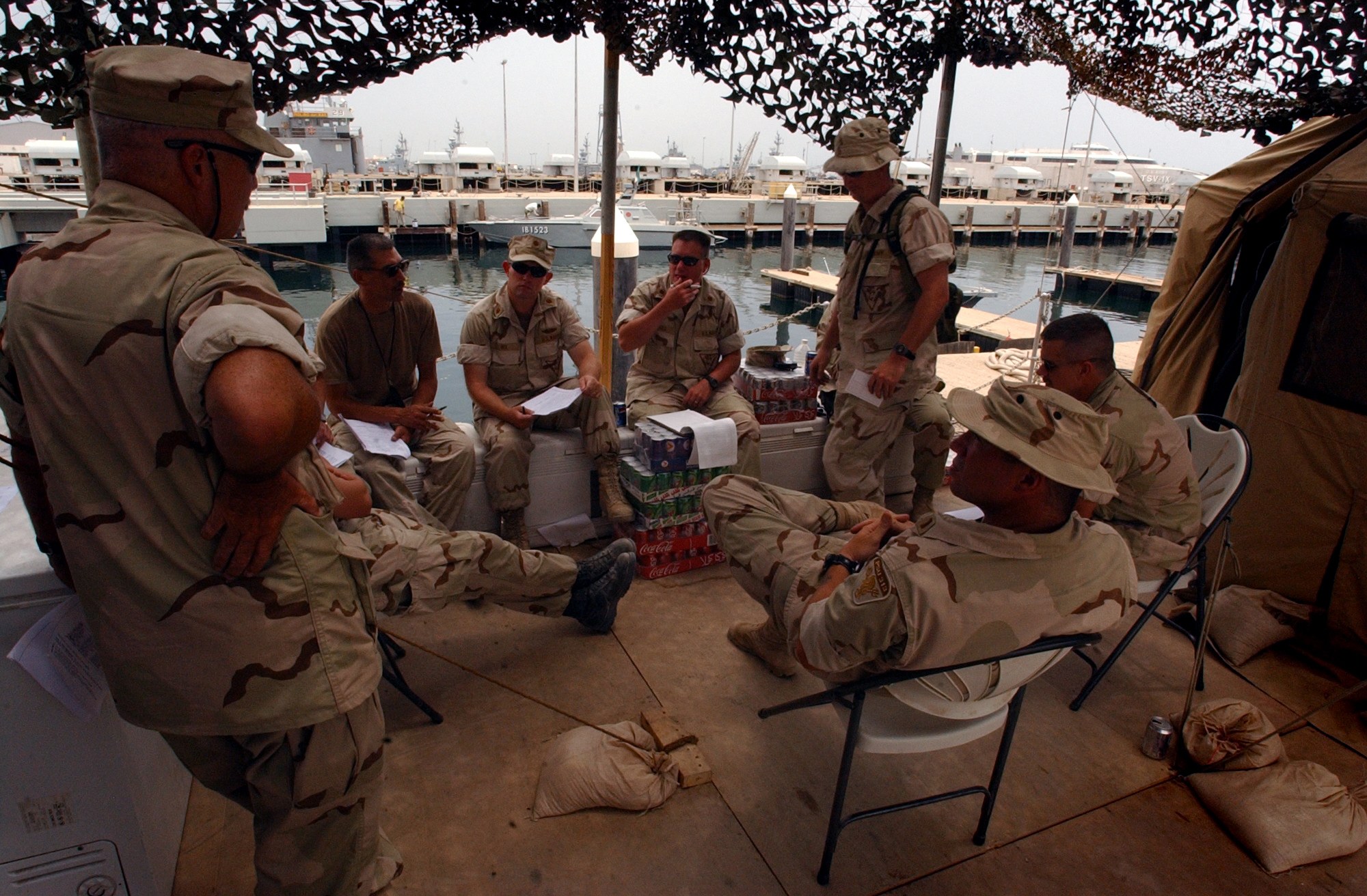 Gruppe von Männern in Mützen und Schwimmbrillen sitzt um einen Tisch auf einem Boot, mit Taschen auf dem Boden und einem Zelt rechts; Boote, Pfähle, Gebäude und ein klarer blauer Himmel im Hintergrund.