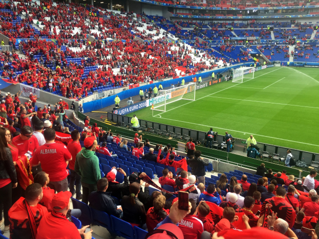 Eine große Menge Menschen in einem Stadion schaut ein Fußballspiel, mit Sitzplätzen und Stehplätzen, Bannern, einem Torpfosten und Deckenleuchten.