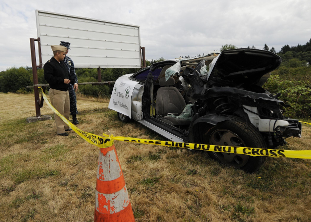 Ein Polizist in Uniform steht neben einem schwer beschädigten Auto am Straßenrand, mit einem Verkehrskegel und einem Band an einem Pfahl in der Nähe und Bäumen und einem bewölkten Himmel im Hintergrund.
