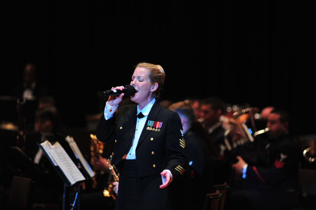 Eine Frau in einer Militäruniform singt in ein Mikrofon auf der Bühne, mit Musikern, die hinter ihr Instrumente spielen und Bücher auf Ständern vorne.
