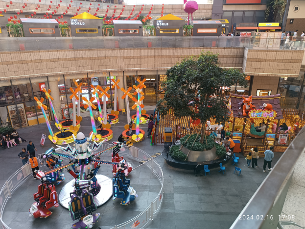 Ein Vergnügungspark innerhalb eines Einkaufszentrums mit verschiedenen Attraktionen, Bäumen, Menschen, Stühlen, Ständen und Gebäuden, mit einem Wasserzeichen in der unteren rechten Ecke.