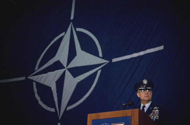 Ein Mann in militärischer Uniform steht an einem Podium mit Mikrofonen vor einer NATO-Flagge, trägt eine Mütze und eine Brille.