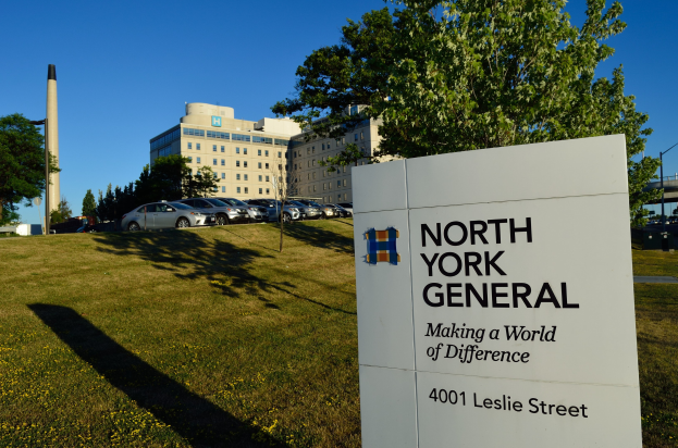 Das North York General Hospital in New York City mit einem Schild im Vordergrund, umgeben von Gras und Blumen, und Fahrzeugen, Bäumen, Gebäuden und Masten im Hintergrund unter einem klaren blauen Himmel.