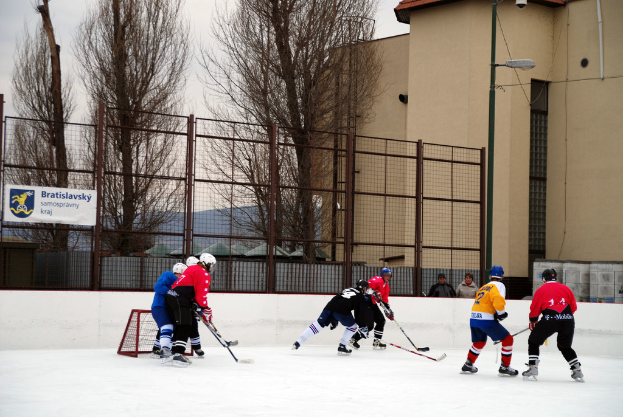 Menschen beim Eishockey auf einem Eisplatz mit Gebäuden, Bäumen, einer Straßenlaterne, einem Namensschild und Zäunen im Hintergrund unter einem Himmel.
