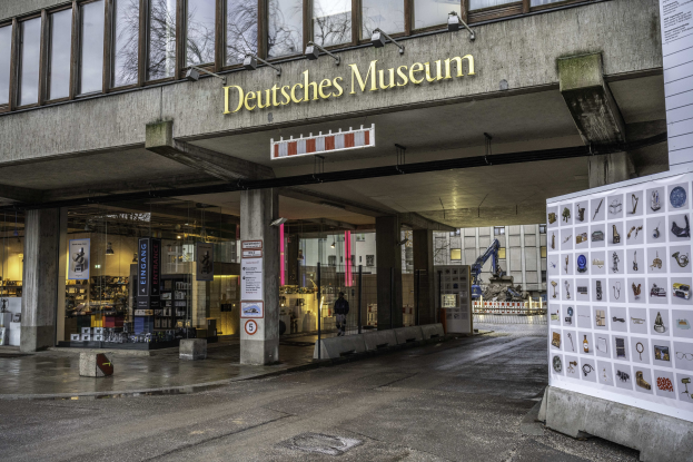 Großes gläsernes Gebäude des Deutschen Museums in Berlin mit einem Namensschild und einer Informationsanzeige, das vor einem Hintergrund von Bäumen und anderen städtischen Strukturen unter dem Himmel zu sehen ist.