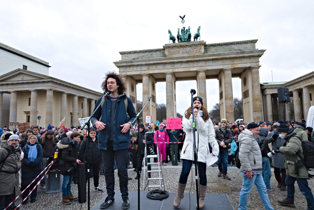 Eine Gruppe von Menschen auf einer Bühne vor dem Brandenburger Tor in Berlin, mit zwei Mikrofonen und einer Kamera in der Hand, einer Leiter im Hintergrund und Gebäuden, Bäumen und einem klaren blauen Himmel im Hintergrund.