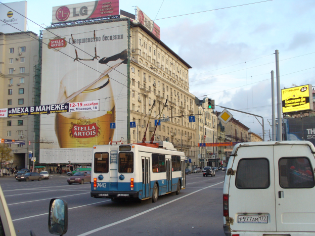 Eine belebte Stadtstraße mit mehreren Fahrzeugen, darunter ein Bus und ein Lieferwagen, umgeben von Gebäuden, Schildern, Strommasten mit Drähten und Verkehrsampeln.