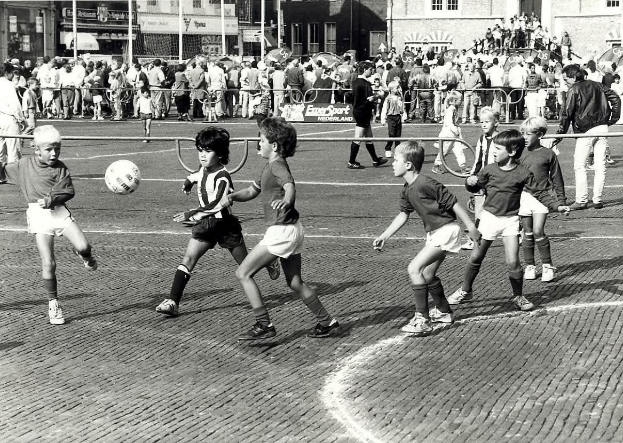 Schwarzes und weißes Bild von jungen Jungs, die Fussball auf der Straße spielen, mit einer Zuschauermenge hinter einem Geländer und Gebäuden im Hintergrund.