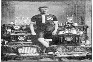 Ein Schwarz-Weiß-Foto eines Mannes in einem Fußballtrikot, der vor einem Tisch voller Trophäen und Pokale steht, mit der Aufschrift "1909-1910 WM-Sieger" unten.