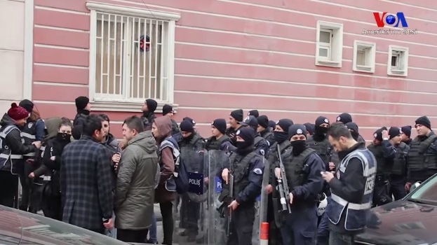 Eine Gruppe von Menschen, einige bewaffnet und mit Möppen, steht vor einem Gebäude mit Fenstern, um gegen eine Regierungsentscheidung zu protestieren, mit Fahrzeugen und einem Verkehrskegel im Vordergrund und einem Wasserzeichen auf dem Bild.