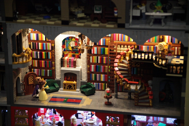 Ein LEGO-Modell eines Bibliotheksinneren mit Bücherregalen, einer Treppe, einem Kronleuchter, einem Kamin, einer Sitzgruppe mit Sofa und Tisch, einem Teppich und verstreuten Spielzeugen.
