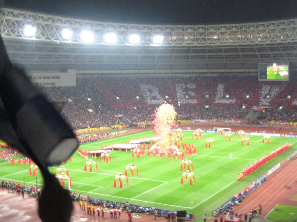 Ein Stadion mit Flutlicht, Eisenstäbe, Zuschauern, stehenden Personen, Werbetafeln, Bildschirme, Überwachungskameras, Fahnen und Dekorationen.