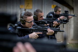 Eine Gruppe von Männern mit Kopfhörern und Gewehren steht vor einer verplankten Wand und nimmt an einem Airsoft-Training teil.
