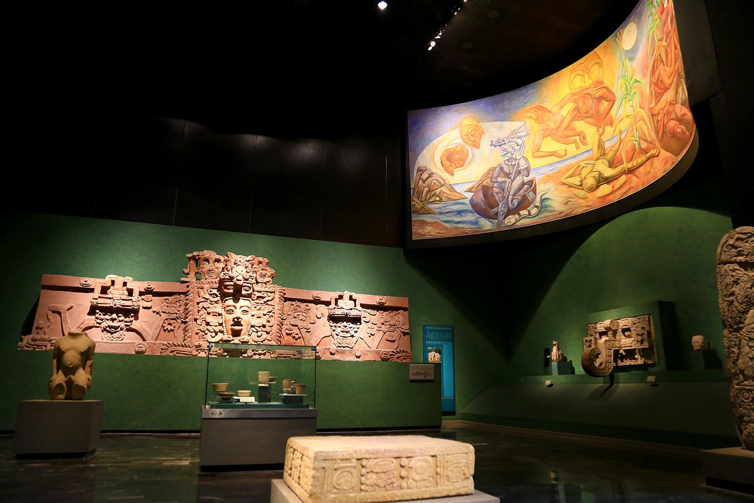 Ausstellungsraum im Anthropologischen Museum von Mexiko-Stadt mit verschiedenen Artefakten, darunter Skulpturen, Tischgegenstände, eine Texttafel, eine Wandmalerei und Deckenbeleuchtung.