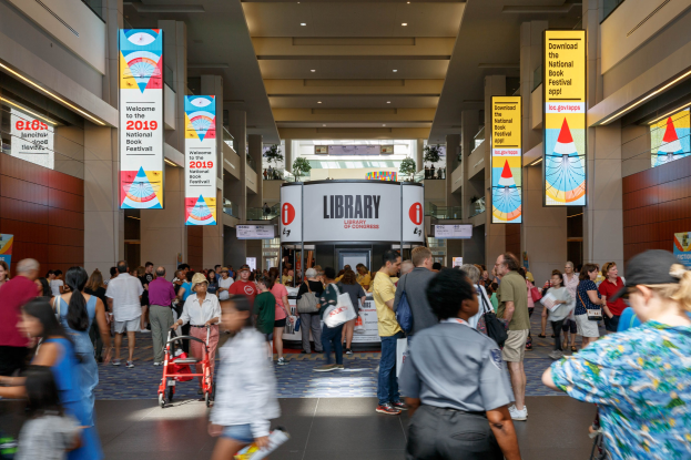 Große Gruppe von Menschen, die durch ein Gebäude mit Bannern an den Wänden und Säulen, Deckenleuchten und einer Tafel im Hintergrund gehen, die mit dem Nationalen Buchfest in Verbindung steht, einige tragen Taschen und einer schiebt einen Kinderwagen.