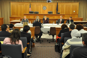 Ein Konferenzraum mit einer Gruppe von Menschen, die auf Stühlen sitzen, zwei ausländischen Flaggen, einem orangen Tisch mit Wasserflaschen und einem Mikrofon.