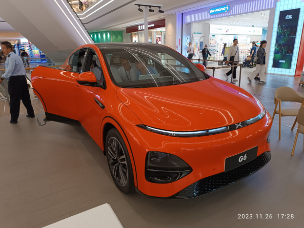 Oranges Elektroauto in einer Ausstellungshalle, umgeben von Menschen, Stühlen, einem Wagen, Geschäften und Namensschildern im Hintergrund.
