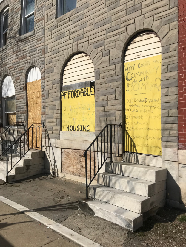 Ein zweistöckiges Gebäude mit vernagelten Fenstern und einem verblichenen "Günstiger Wohnraum"-Schild, das über eine Treppe mit Geländern erreichbar ist und neben einer Straße steht.