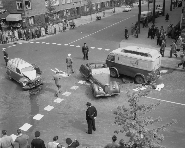 Schwarz-weiß-Szene eines Autounfalls an einer Straßenkreuzung mit mehreren Fahrzeugen, darunter ein Van, und einer Gruppe von Schaulustigen; Gebäude, Bäume, Strommasten und Texttafeln sind im Hintergrund zu sehen.