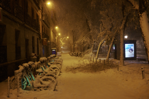 Eine verschneite Straße bei Nacht mit parkenden Fahrrädern, Bäumen, Gebäuden, Straßenlaternen, Pfählen, Brettern und einem Bildschirm im Hintergrund, der eine friedliche Winterszene schafft.