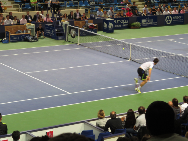 Ein Mann spielt Tennis auf einem Court mit einem Ball in der Luft, der einen Schläger hält, vor einer sitzenden Menge, mit einem Netz, Bannern und Stufen im Hintergrund.