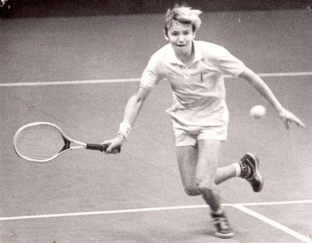Ein junger Mann in weißer Tennisbekleidung schlägt mit einem Schläger einen Ball auf einem Tennisplatz, mit entschlossener Miene.