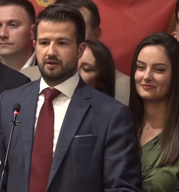 Ein Mann im Anzug spricht an einem Rednerpult mit einem Mikrofon und blickt auf eine Menge hinter einem Banner, wahrscheinlich über ein Regierungssozialmedienverbot.