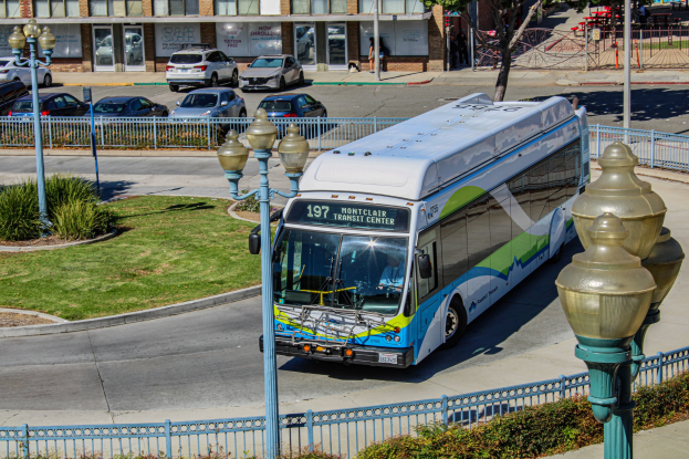 Ein Bus fährt auf einer Straße mit Laternen, Geländern, Grünflächen und Gebäuden im Hintergrund, mit Fußgängern auf dem Gehweg.