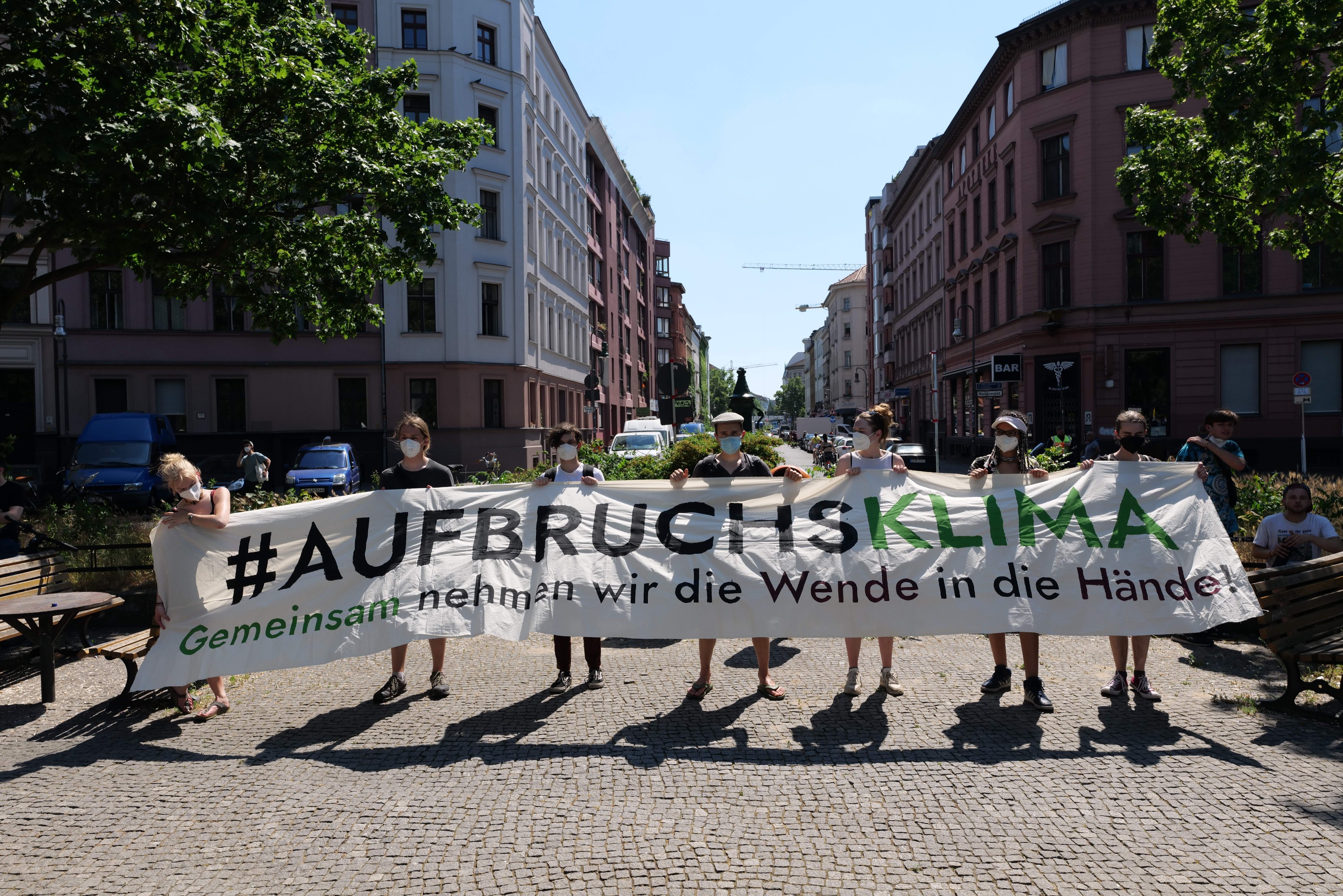 Eine Gruppe von Menschen mit Masken, die ein Banner mit der Aufschrift 'Aufbruchsklima' während einer Klimaprotest in Berlin, Deutschland, halten, umgeben von urbanen Elementen wie Bänken, Tischen und Fahrzeugen.