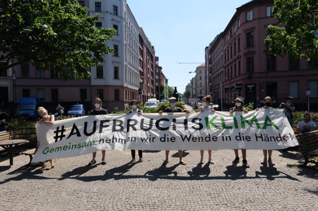 Eine Gruppe von Menschen mit Masken, die ein Banner mit der Aufschrift 'Aufbruchsklima' während einer Klimaprotest in Berlin, Deutschland, halten, umgeben von urbanen Elementen wie Bänken, Tischen und Fahrzeugen.