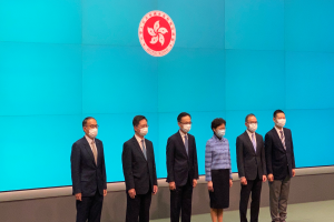 Eine Gruppe von Menschen mit Masken steht vor einer blauen Wand mit einem Logo und nimmt an einer Pressekonferenz in Hong Kong teil.