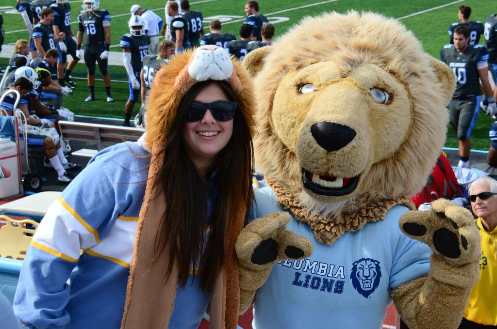 Eine Frau steht neben einer Person in einem Löwenkostüm, mit Spielern auf dem Boden dahinter und Zuschauern auf Bänken und Stühlen links.