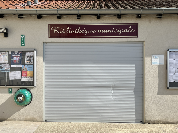 Gebäude der städtischen Bibliothek mit einem "Bibliothèque municipale"-Schild, einer zentralen Jalousie, einer Texttafel, Plakaten an der linken Wand, beleuchteter Bereich und einem Weg, der zur Eingangstür führt.