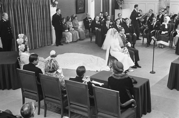 Schwarzes und weißes Foto einer Hochzeitszeremonie mit sitzenden und stehenden Menschen, ein Mann spricht in ein Mikrofon und eine Frau hält einen Blumenstrauß, mit einem Podium, Blumengestecken, einem Vorhang, einem gerahmten Foto an der Wand und einer Tür im Hintergrund.