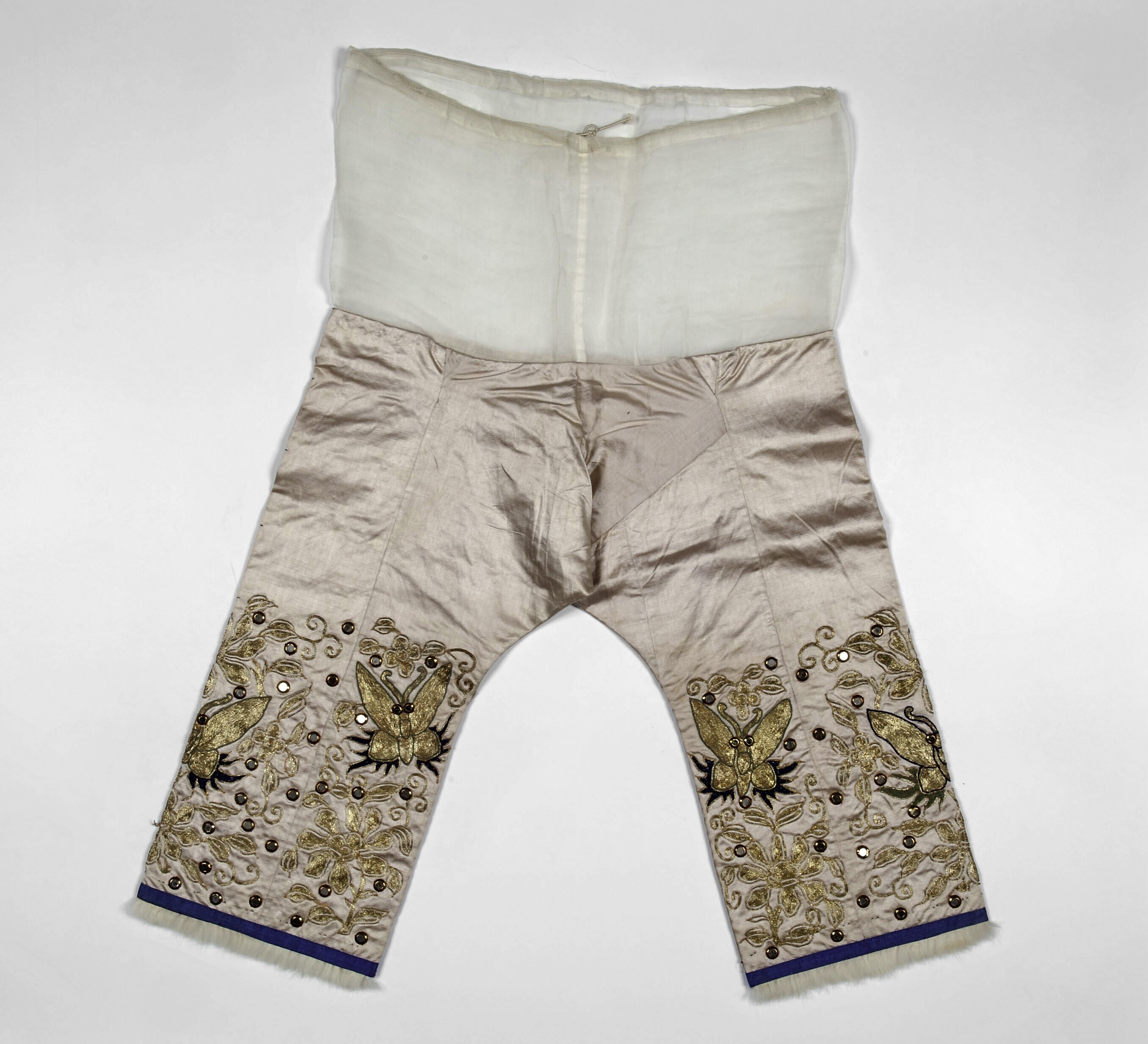 Ein Paar weiße, hochtaillierte, weite Hosen mit aufwändiger Goldstickerei, vor einem weißen Hintergrund.