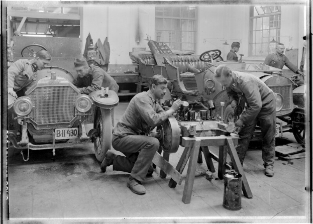 Ein Schwarz-Weiß-Bild von mehreren uniformierten Männern, die einen Wagen in einer Garage reparieren, umgeben von Werkzeugen und Equipment auf einem zentralen Tisch.