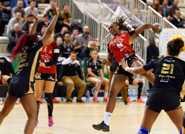 Eine Gruppe von Frauen, die Volleyball auf einem Feld mit einem Ball in der Mitte spielen, während Zuschauer im Hintergrund auf Stühlen sitzen, Geländer und Flaschen auf dem Boden.