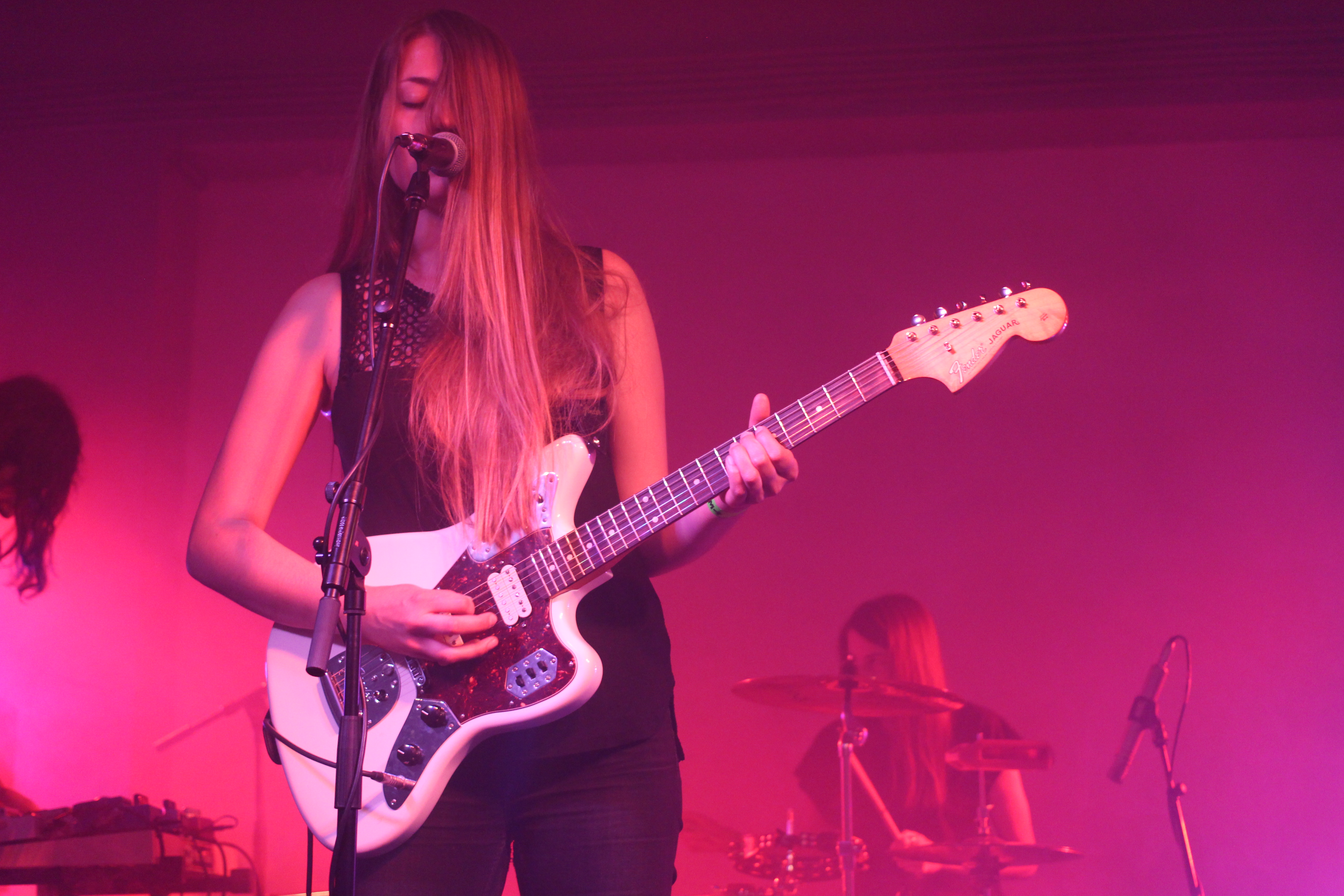 Eine Frau spielt Gitarre auf der Bühne bei einem Konzert, mit einem Mikrofon vor ihr und einer anderen Person, die ein Musikinstrument spielt, links von ihr.
