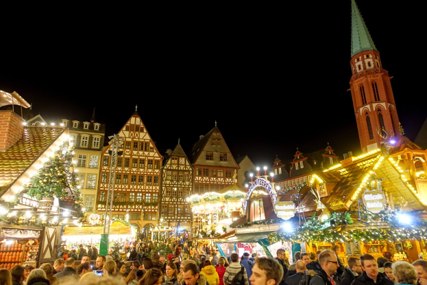Ein geschäftiger Weihnachtsmarkt in Nürnberg, Deutschland mit Menschen um dekorierte Stände, festliche Lichter, Gebäude mit Fenstern, einen Kirchturm und einen dunklen Himmel.