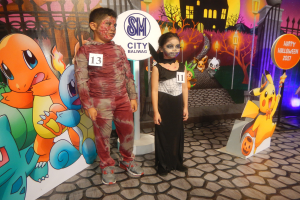 Zwei Kinder in Halloween-Kostümen stehen nebeneinander vor einer Wand mit einem Gemälde eines Gebäudes, Bäumen, Laternenmasten und einem Zaun sowie Schildern mit Text und Tierbildern und festlicher Beleuchtung.