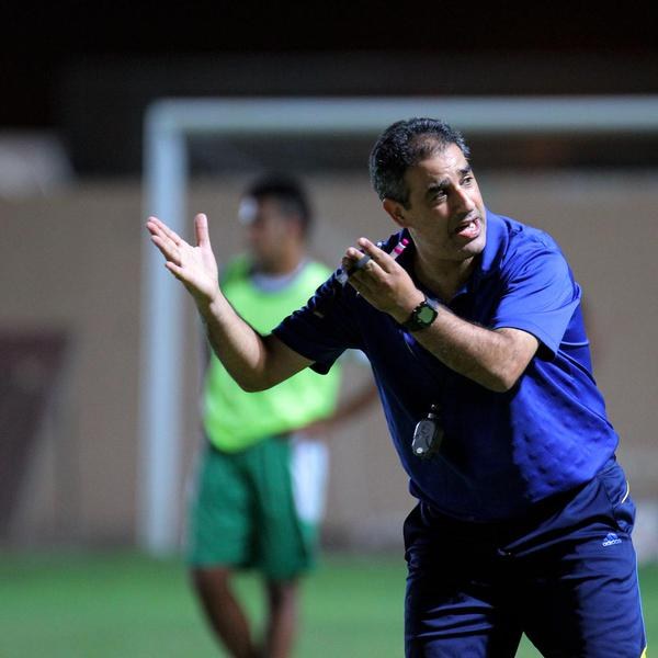 Ein Mann in einem blauen T-Shirt und Hose steht auf einem Fußballfeld und hält ein Mikrofon in der Hand, während er in Richtung der Spieler gestikuliert. Im Hintergrund ist eine Person in einem grünen Kleid, ein Torpfosten und eine Wand zu sehen.