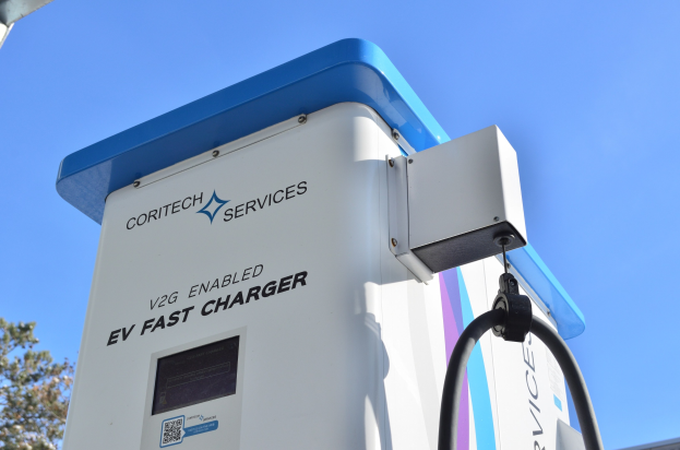 Elektrofahrzeug-Ladestation mit der Beschriftung "V2G Fast Charger" mit B├Ąumen und einem Geb├Ąude im Hintergrund bei klarem blauem Himmel.