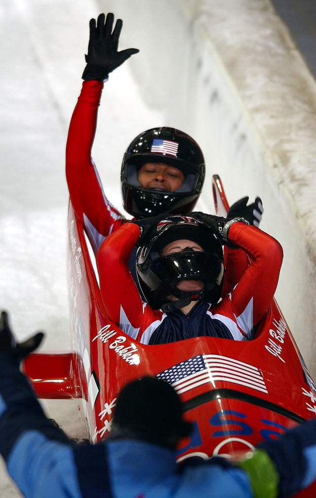 Zwei Bobsleighs beim olympischen Wettbewerb, eines mit Helm und Handschuhen, mit einer Person und einer Wand im Hintergrund.
