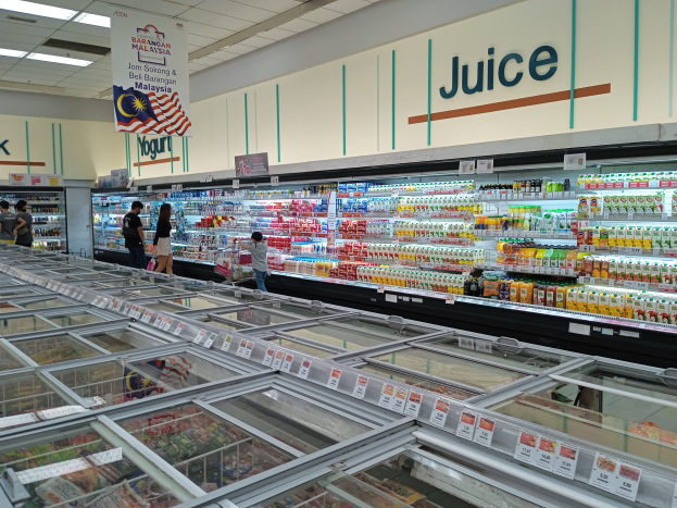 Ein hell erleuchteter Gang in einem Supermarkt mit einer großen Auswahl an Getränken, frischem Obst und Gemüse sowie anderen Lebensmitteln, mit einigen Kunden, die durch die Gänge schlendern, und einer beleuchteten Tafel an der Decke.