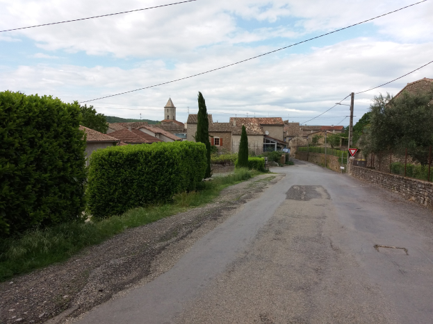 Kleiner Ort mit einer von Bäumen und Büschen gesäumten Straße, die zu Gebäuden im Hintergrund führt, einschließlich eines Schildes auf der rechten Seite.