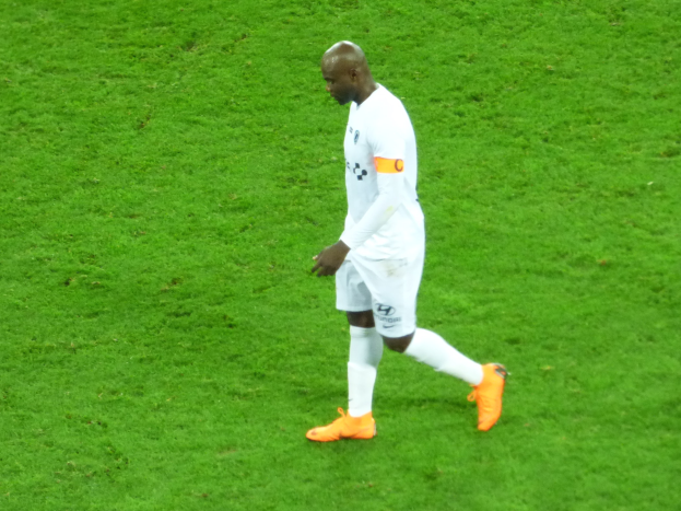 Ein Fußballspieler in einem weißen T-Shirt, weißen Shorts und orangefarbenen Schuhen läuft über ein saftiges Grün.