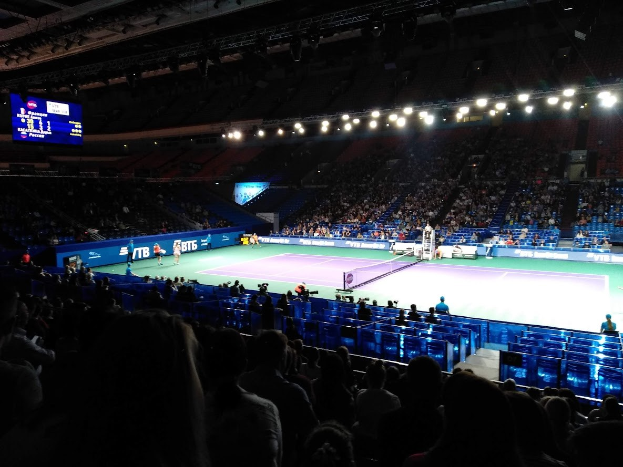 Ein Tennis-Spiel in einer großen Arena mit Zuschauern auf Stühlen, ein Netz teilt den Court, einige stehende Personen, ein Bildschirm mit Text auf der linken Seite und Überdachungsbeleuchtung.